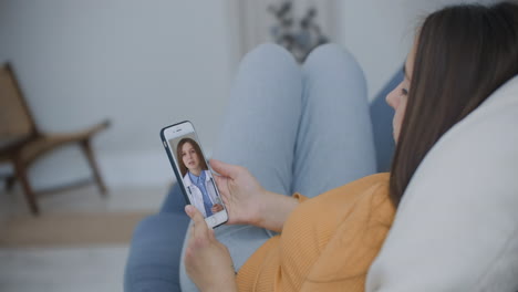 A-woman-lying-on-the-couch-talking-to-a-woman-doctor-on-video-using-a-smartphone.-Remote-medical-consultation-with-a-doctor-via-mobile-phone